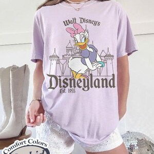 Puede incluir: Una camiseta lila con un dise&ntilde;o de Daisy Duck frente al castillo de Disneyland. El texto en la camiseta dice "Walt Disney's Disneyland Est. 1955". La camiseta est&aacute; etiquetada como "Comfort Colors Orchid".