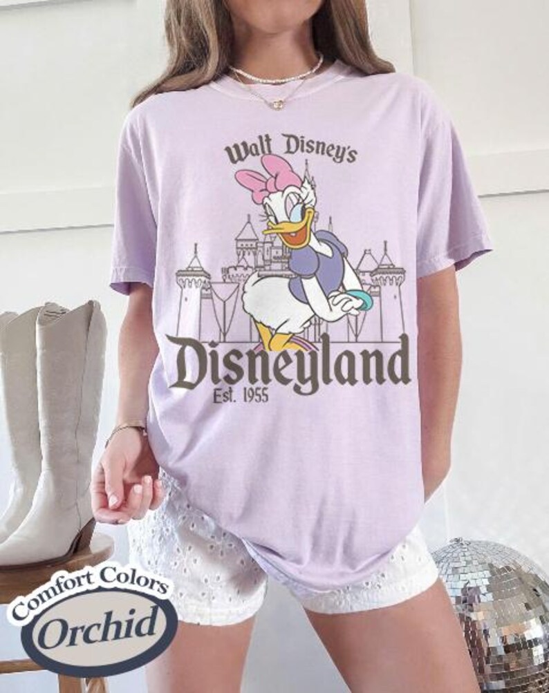 Puede incluir: Una camiseta lila con un dise&ntilde;o de Daisy Duck frente al castillo de Disneyland. El texto en la camiseta dice "Walt Disney's Disneyland Est. 1955". La camiseta est&aacute; etiquetada como "Comfort Colors Orchid".