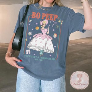 May include: Blue t-shirt featuring a Bo Peep graphic from Toy Story. The design includes the text "Bo Peep" and "You've Got a Friend in Me Est. 1997". The graphic is framed with a colorful border and includes stars and other Toy Story characters.