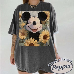 Puede incluir: Camiseta gris oscuro con un dise&ntilde;o de Mickey Mouse sonriendo rodeado de girasoles. El dise&ntilde;o est&aacute; centrado en la parte delantera. La camiseta tiene mangas cortas y un corte holgado. El texto "Comfort Colors Pepper" est&aacute; impreso en la parte inferior derecha.