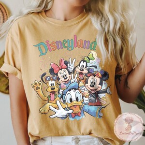 May include: Mustard yellow t-shirt featuring a colorful graphic of Disney characters, including Mickey and Minnie Mouse, Donald Duck, and Goofy. The word "Disneyland" is written across the top in a rainbow font. The characters are holding ice cream cones and posing for a photo.