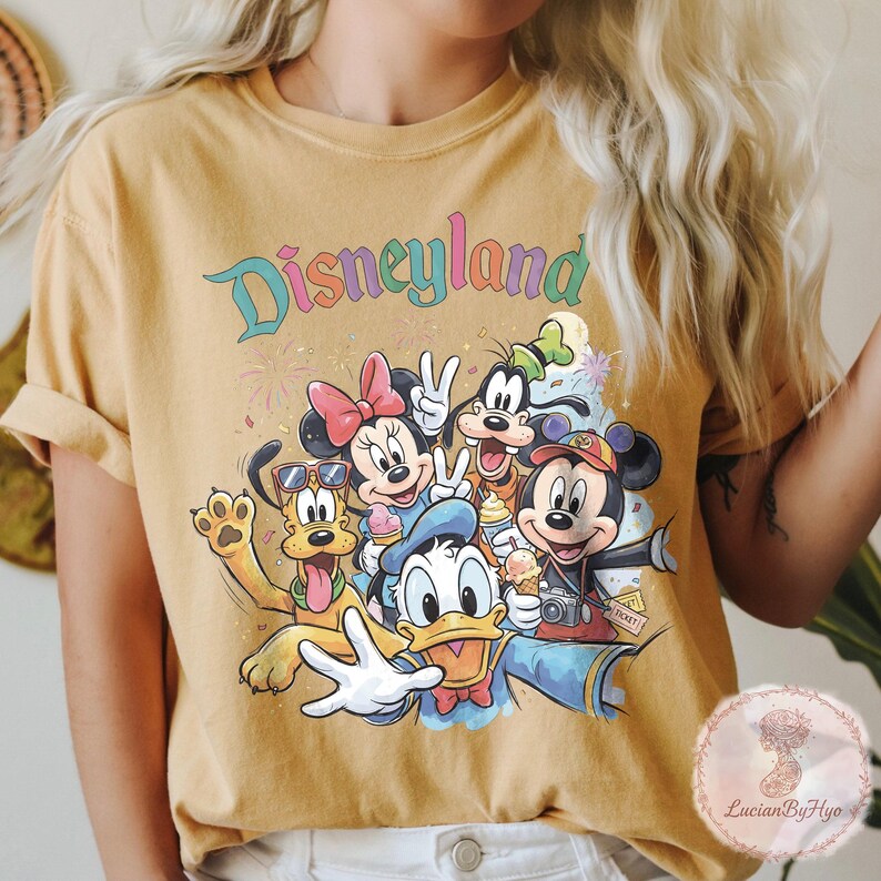 May include: Mustard yellow t-shirt featuring a colorful graphic of Disney characters, including Mickey and Minnie Mouse, Donald Duck, and Goofy. The word "Disneyland" is written across the top in a rainbow font. The characters are holding ice cream cones and posing for a photo.
