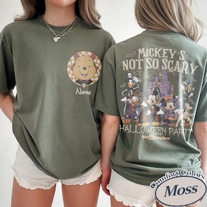 May include: Olive green t-shirts with Halloween-themed graphics. One shirt features Winnie the Pooh in a costume with the word "Name" below. The other shirt displays "Mickey's Not So Scary Halloween Party" with Disney characters and a castle.