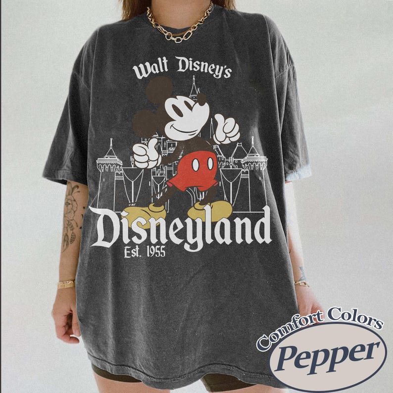 Puede incluir: Camiseta gris oscuro con Mickey Mouse y el texto "Walt Disney's Disneyland Est. 1955". El dise&ntilde;o muestra a Mickey Mouse con los brazos levantados y un castillo. La camiseta tiene un aspecto vintage.