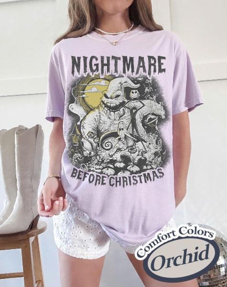 May include: A light purple t-shirt featuring a graphic from "The Nightmare Before Christmas." The design includes the text "NIGHTMARE BEFORE CHRISTMAS" and characters from the movie. The shirt is labeled "Comfort Colors" and "Orchid."