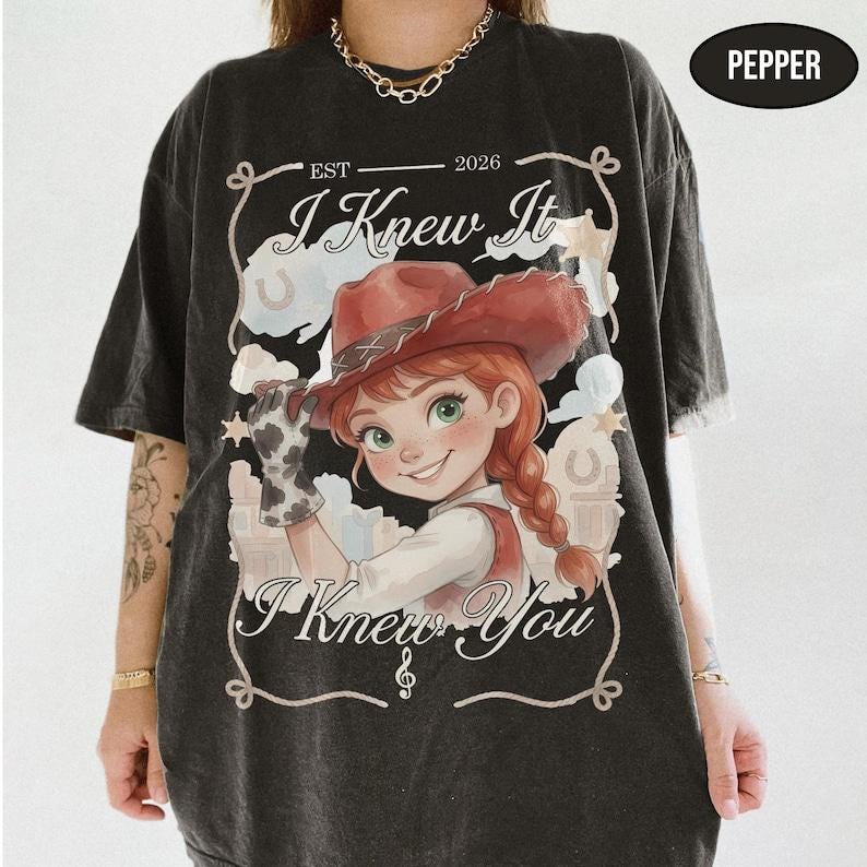 May include: A charcoal gray t-shirt featuring a graphic of a cartoon cowgirl with red hair in a braid and a cowboy hat. The text reads "EST 2026", "I Knew It", and "I Knew You" with a musical note. A black oval tag in the corner says "PEPPER".