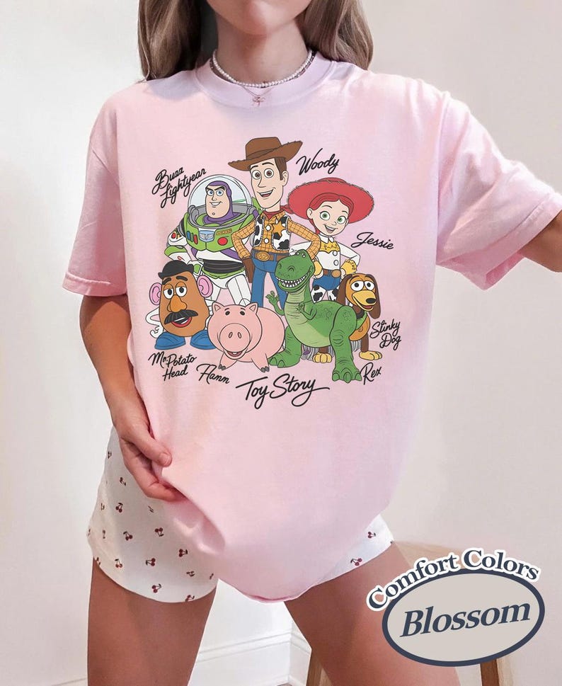 May include: A pale pink t-shirt with a graphic of characters from the animated film "Toy Story." The design includes Woody, Buzz Lightyear, Jessie, and other characters. The text "Toy Story" is also visible.