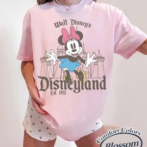 Puede incluir: Camiseta rosa claro con un dise&ntilde;o de Minnie Mouse frente a un castillo y el texto "Walt Disney's Disneyland Est. 1955". Pertenece a la colecci&oacute;n Comfort Colors Blossom.