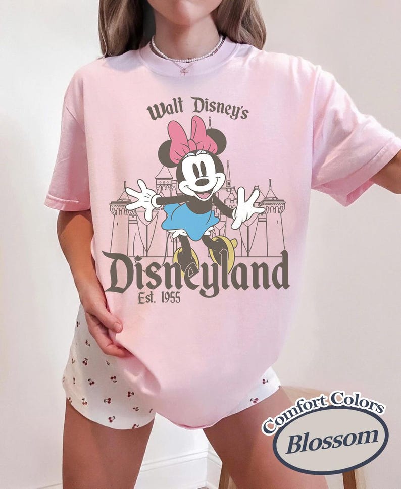 Puede incluir: Camiseta rosa claro con un dise&ntilde;o de Minnie Mouse frente a un castillo y el texto "Walt Disney's Disneyland Est. 1955". Pertenece a la colecci&oacute;n Comfort Colors Blossom.