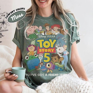 May include: A moss green short-sleeve t-shirt featuring a graphic print of various Toy Story characters, the Disney Pixar logo, the text Toy Story 5, and the phrase You've Got A Friend In Me. A sticker in the top left corner reads Comfort Colors Moss.