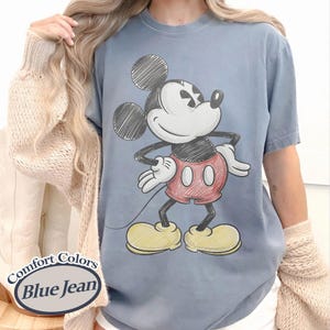 May include: Blue jean Comfort Colors t-shirt featuring a sketched Mickey Mouse graphic. The cartoon character is in red shorts, yellow shoes, and white gloves. The shirt has the words "Comfort Colors" and "Blue Jean" on the bottom left.