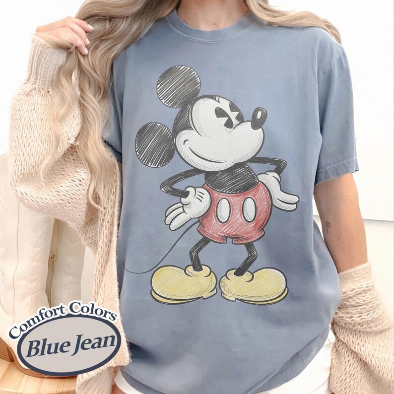 May include: Blue jean Comfort Colors t-shirt featuring a sketched Mickey Mouse graphic. The cartoon character is in red shorts, yellow shoes, and white gloves. The shirt has the words "Comfort Colors" and "Blue Jean" on the bottom left.