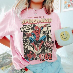 May include: A light pink t-shirt featuring a graphic print of Spider-Man in a dynamic pose. The text reads THE AMAZING SPIDER-MAN, NEW YORK, SWING, SPRING INTO ACTION, and 1962. The design includes a comic book style illustration of the character.