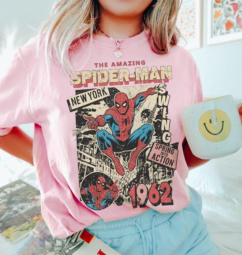 May include: A light pink t-shirt featuring a graphic print of Spider-Man in a dynamic pose. The text reads THE AMAZING SPIDER-MAN, NEW YORK, SWING, SPRING INTO ACTION, and 1962. The design includes a comic book style illustration of the character.