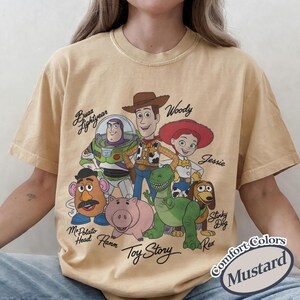 May include: Mustard-coloured t-shirt with a graphic of Toy Story characters, including Woody, Buzz Lightyear, and Jessie. The shirt has the words "Toy Story" and character names printed on it. The shirt is labelled "Comfort Colors".