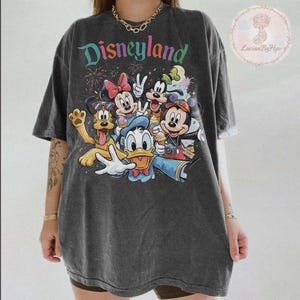 May include: Dark gray t-shirt featuring the word "Disneyland" in colorful text above a graphic of Disney characters including Mickey Mouse, Minnie Mouse, Donald Duck, Goofy, and Pluto. The shirt has a vintage, worn look.