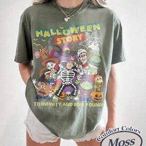 May include: Moss green Comfort Colors t-shirt with a Halloween-themed graphic. The graphic features various cartoon characters, the words "HALLOWEEN STORY", and the phrase "TO INFINITY AND BOO YOUND!"