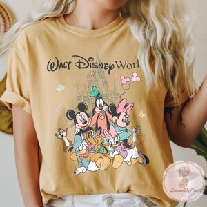 May include: Mustard yellow t-shirt featuring a graphic of Disney characters including Mickey Mouse, Minnie Mouse, Goofy, Donald Duck, and Daisy Duck. The words "Walt Disney World" are printed above the characters, with a castle in the background.