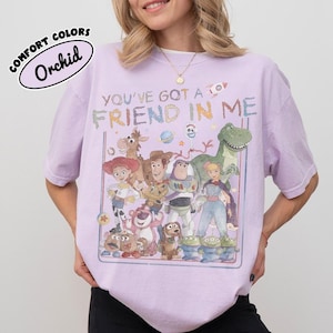 Pu&ograve; includere: Una t-shirt a maniche corte color viola chiaro con una stampa grafica di vari personaggi animati e il testo "YOU'VE GOT A FRIEND IN ME". Il design include un cowboy, un ranger spaziale, una cowgirl, un dinosauro e altre figure in un riquadro.