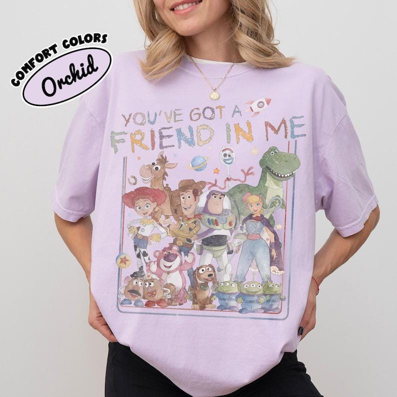 Pu&ograve; includere: Una t-shirt a maniche corte color viola chiaro con una stampa grafica di vari personaggi animati e il testo "YOU'VE GOT A FRIEND IN ME". Il design include un cowboy, un ranger spaziale, una cowgirl, un dinosauro e altre figure in un riquadro.