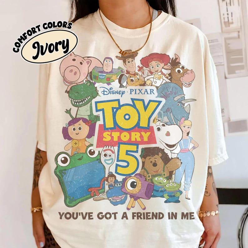 May include: An ivory t-shirt featuring a graphic print of various Toy Story characters including Woody, Buzz Lightyear, and Jessie. The text reads Disney Pixar Toy Story 5 and You've Got A Friend In Me. A Comfort Colors Ivory label is visible in the corner.