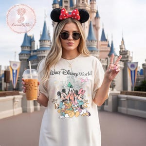 May include: Cream-colored t-shirt featuring a graphic of Disney characters, including Mickey Mouse, Goofy, and Minnie Mouse, with the text "Walt Disney World." The person is wearing a Minnie Mouse ear headband with a red bow and sunglasses.