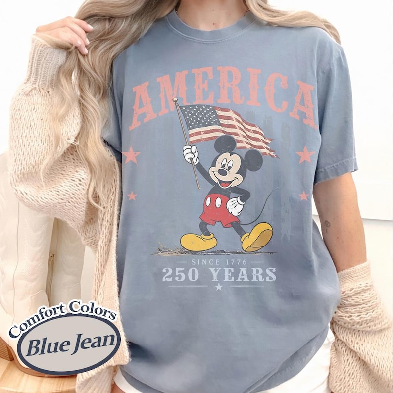 Pu&ograve; includere: T-shirt blu jeans Comfort Colors con Topolino che tiene una bandiera americana. La maglietta ha la scritta "AMERICA" sopra la grafica e "250 YEARS SINCE 1776" sotto. Stelle rosa sulla maglietta.