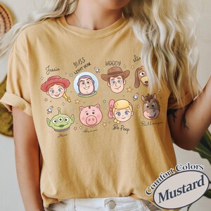 May include: Mustard-colored t-shirt featuring cartoon illustrations of characters from the Toy Story movie franchise. The shirt includes characters like Woody, Buzz Lightyear, and Jessie. The text "Comfort Colors" and "Mustard" are also visible.