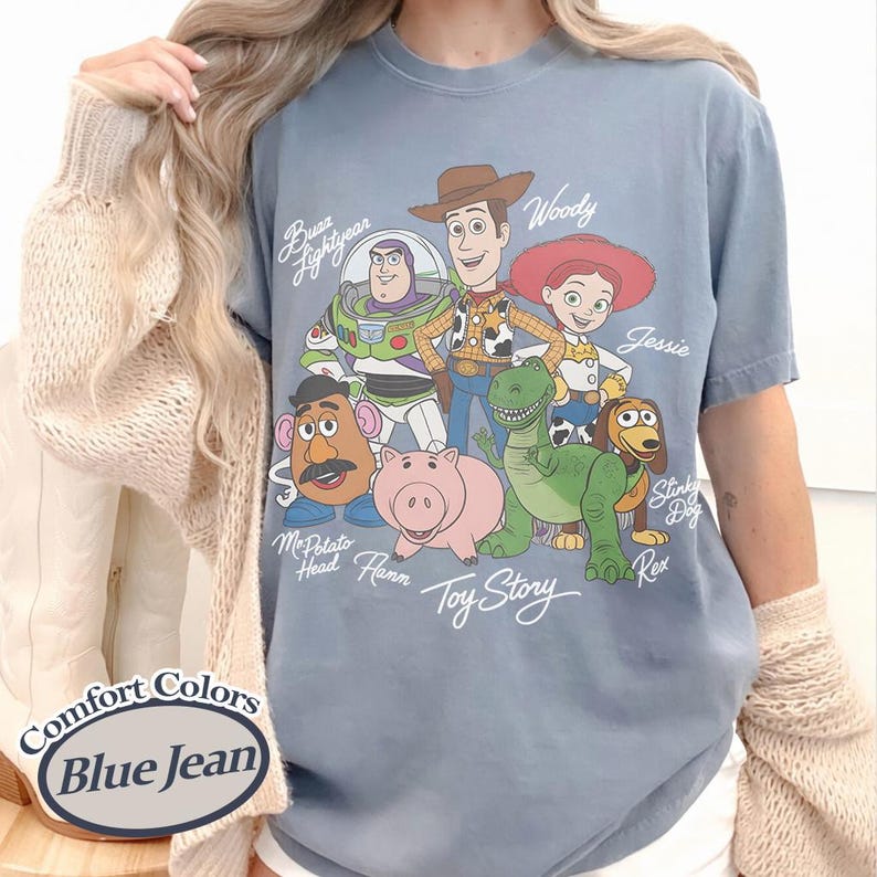 May include: Blue jean Comfort Colors t-shirt with a graphic of Toy Story characters, including Woody, Buzz Lightyear, Jessie, and Rex. The shirt has a crew neck and short sleeves. The text "Toy Story" is printed below the characters.