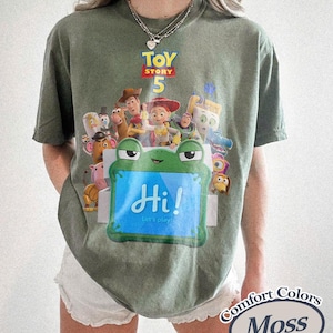 May include: A moss green short-sleeve t-shirt featuring a graphic print of Toy Story 5 characters and a green frog-shaped box with the text Hi! Let's play! The shirt has a crew neckline and a relaxed fit, with a Comfort Colors Moss label visible.