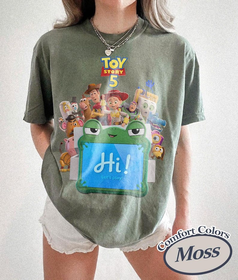 May include: A moss green short-sleeve t-shirt featuring a graphic print of Toy Story 5 characters and a green frog-shaped box with the text Hi! Let's play! The shirt has a crew neckline and a relaxed fit, with a Comfort Colors Moss label visible.