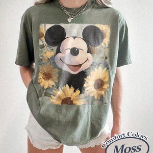 Puede incluir: Camiseta verde oliva con un dise&ntilde;o de Mickey Mouse sonriente rodeado de girasoles. La camiseta es de la marca Comfort Colors y tiene un corte holgado. El gr&aacute;fico es una colorida ilustraci&oacute;n del ic&oacute;nico personaje de dibujos animados.