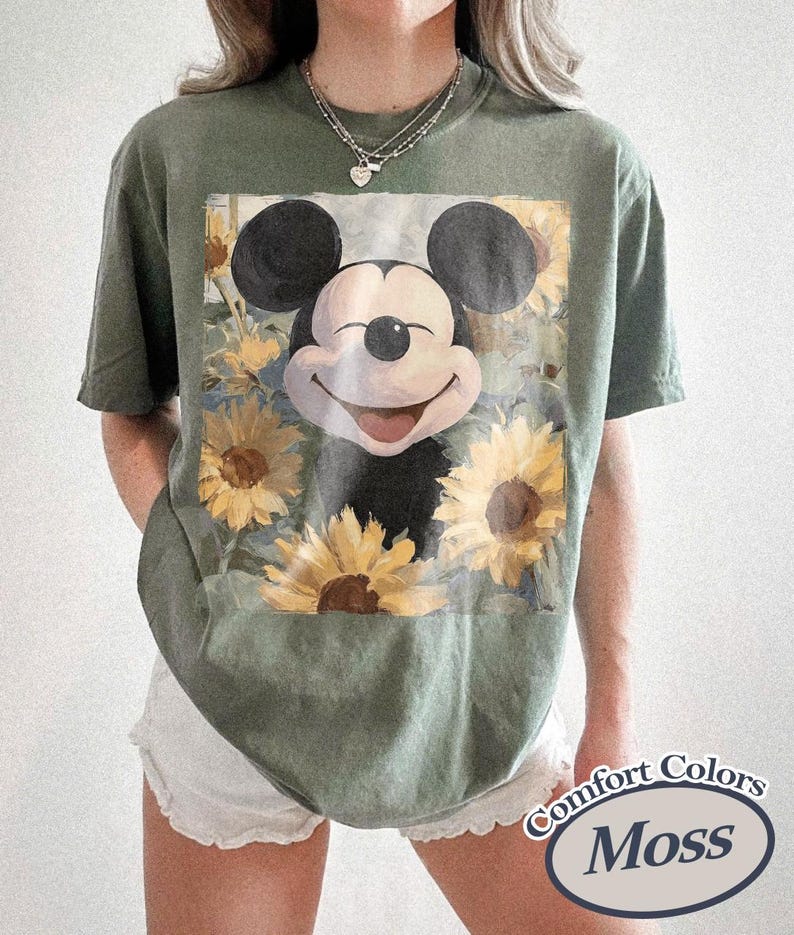 Puede incluir: Camiseta verde oliva con un dise&ntilde;o de Mickey Mouse sonriente rodeado de girasoles. La camiseta es de la marca Comfort Colors y tiene un corte holgado. El gr&aacute;fico es una colorida ilustraci&oacute;n del ic&oacute;nico personaje de dibujos animados.