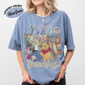 May include: A blue jean coloured t-shirt featuring a graphic print of Winnie the Pooh, Tigger, Piglet, Eeyore, and Rabbit in front of a castle. The text "Disneyland Est. 1955" is printed below the characters. A "Comfort Colors Blue Jean" logo is visible.