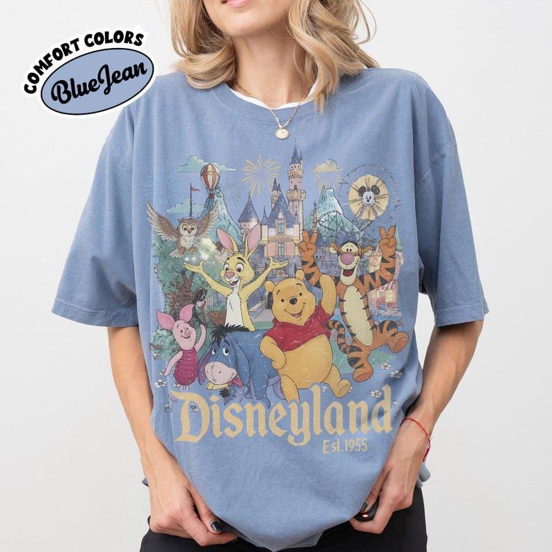 May include: A blue jean coloured t-shirt featuring a graphic print of Winnie the Pooh, Tigger, Piglet, Eeyore, and Rabbit in front of a castle. The text "Disneyland Est. 1955" is printed below the characters. A "Comfort Colors Blue Jean" logo is visible.