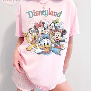 May include: Pink t-shirt with a Disneyland graphic featuring Mickey Mouse, Minnie Mouse, Donald Duck, Goofy, and Pluto. The word "Disneyland" is written in colorful letters above the characters. The shirt is paired with pink and white checkered shorts.
