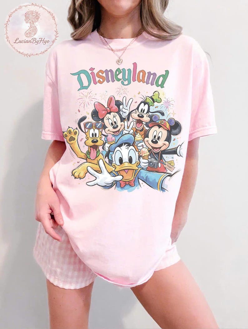 May include: Pink t-shirt with a Disneyland graphic featuring Mickey Mouse, Minnie Mouse, Donald Duck, Goofy, and Pluto. The word "Disneyland" is written in colorful letters above the characters. The shirt is paired with pink and white checkered shorts.
