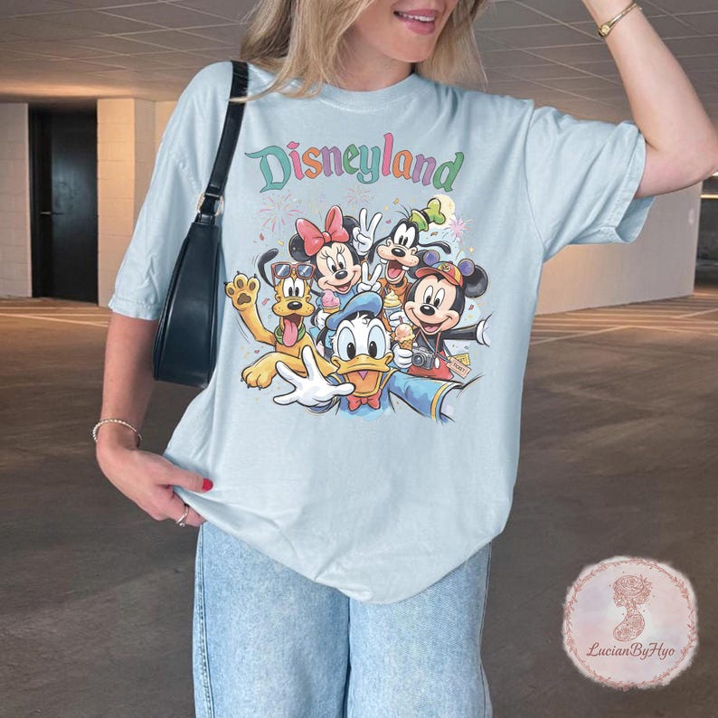 May include: Light blue t-shirt with the word "Disneyland" in a colorful font. The shirt features a graphic of Disney characters including Mickey Mouse, Minnie Mouse, Donald Duck, Goofy, and Pluto. The characters are smiling and holding ice cream cones.