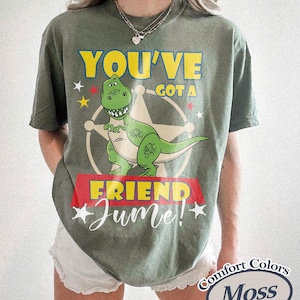 Op de afbeelding: Mosgroen t-shirt met een afbeelding van een groene dinosaurus, de tekst "YOU'VE GOT A FRIEND" in geel en "Jume!" in witte letters. Het shirt heeft een vintage uitstraling.