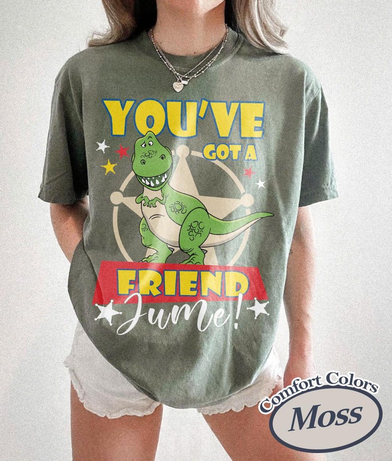 Op de afbeelding: Mosgroen t-shirt met een afbeelding van een groene dinosaurus, de tekst "YOU'VE GOT A FRIEND" in geel en "Jume!" in witte letters. Het shirt heeft een vintage uitstraling.