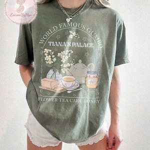 May include: Olive green t-shirt with a graphic featuring the text "WORLD FAMOUS GUMBO TIANA'S PALACE". The design includes illustrations of flowers, a teapot, a teacup, honey, and cake. The words "FLOWER TEA CAKE HONEY" are also printed on the shirt.