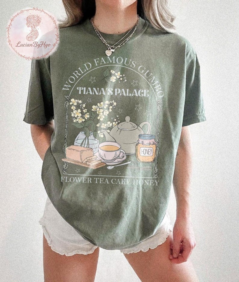 May include: Olive green t-shirt with a graphic featuring the text "WORLD FAMOUS GUMBO TIANA'S PALACE". The design includes illustrations of flowers, a teapot, a teacup, honey, and cake. The words "FLOWER TEA CAKE HONEY" are also printed on the shirt.