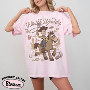 May include: A light pink t-shirt featuring a graphic print of the character Sheriff Woody riding a brown horse. The design includes the text Sheriff Woody, a lasso, cacti, skulls, and stars in a desert-themed illustration on the front of the shirt.