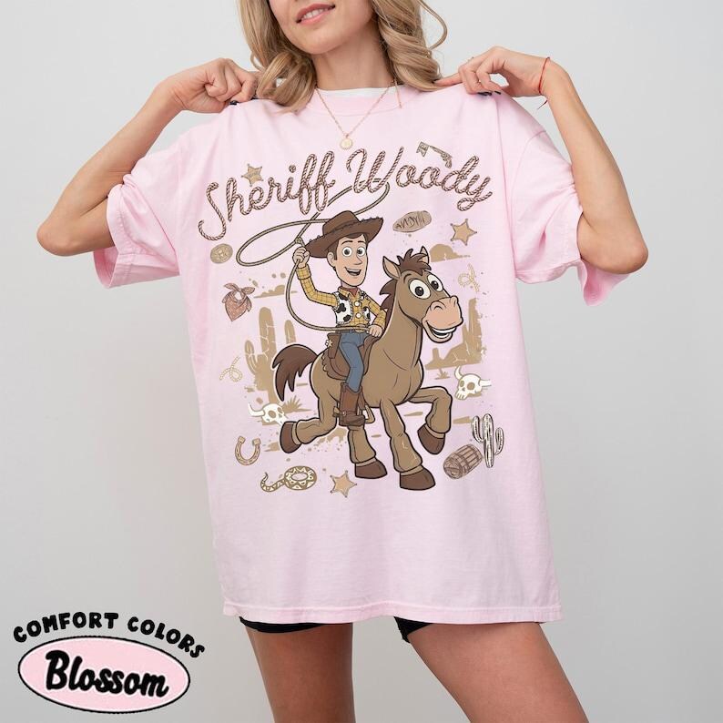 May include: A light pink t-shirt featuring a graphic print of the character Sheriff Woody riding a brown horse. The design includes the text Sheriff Woody, a lasso, cacti, skulls, and stars in a desert-themed illustration on the front of the shirt.