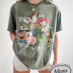 May include: A moss green t-shirt with a graphic of characters from the animated film "Toy Story." The design includes Woody, Buzz Lightyear, Jessie, and other characters. The shirt is labelled "Comfort Colors Moss."