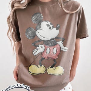 May include: Brown t-shirt featuring a sketch-style Mickey Mouse graphic. The cartoon character is in black, red, and yellow. The shirt has the words "Comfort Colors Espresso" printed on it.
