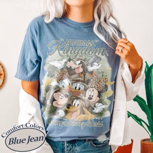 May include: Blue jean colored t-shirt featuring a graphic of Disney characters, including Mickey and Minnie Mouse, Donald and Daisy Duck, Goofy, and Pluto. The shirt has the words "Animal Kingdom" and "Walt Disney World" printed on it.