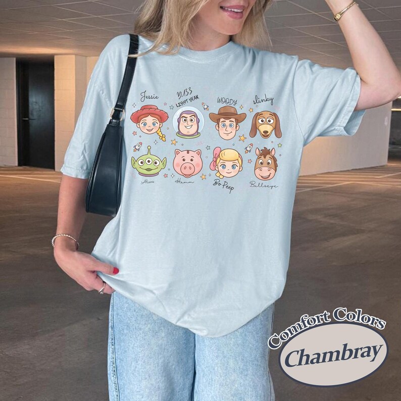 May include: Light blue Comfort Colors Chambray t-shirt featuring cartoon character faces from a popular animated movie. The shirt includes the names Jessie, Buzz Lightyear, Woody, Slinky, Alien, Hamm, Bo Peep, and Bullseye.