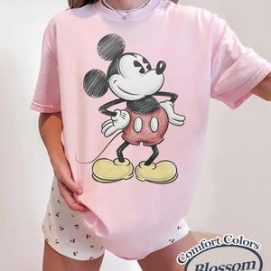 May include: A light pink t-shirt featuring a sketch-style Mickey Mouse graphic. The cartoon character is depicted in black, red, and yellow. The shirt has short sleeves and a relaxed fit. The text "Comfort Colors Blossom" is in the bottom right corner.