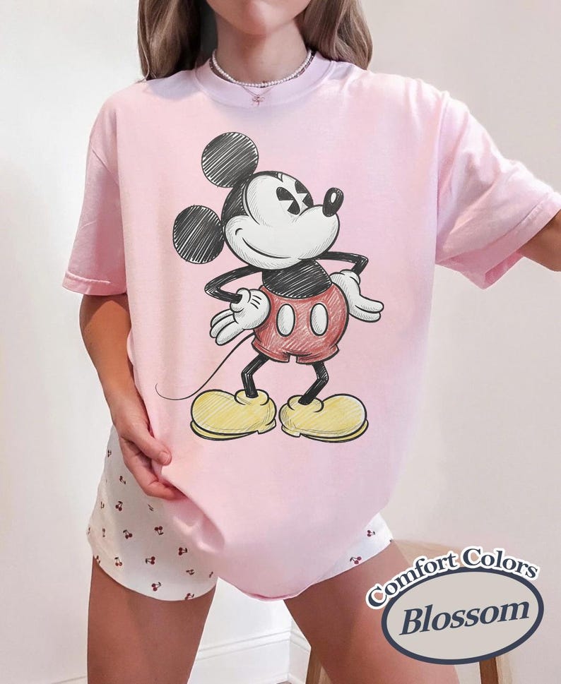 May include: A light pink t-shirt featuring a sketch-style Mickey Mouse graphic. The cartoon character is depicted in black, red, and yellow. The shirt has short sleeves and a relaxed fit. The text "Comfort Colors Blossom" is in the bottom right corner.